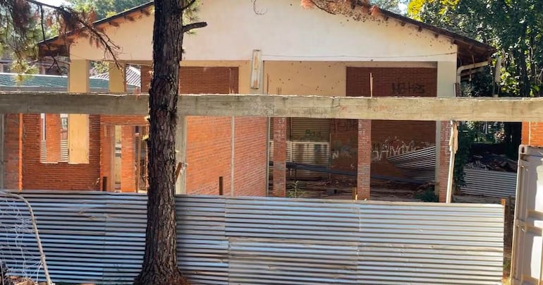 Construcción en estado de abandono rodeada de árboles, con valla de metal y techo deteriorado.