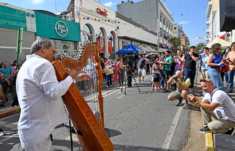 Músico de espaldas con camisa blanca tocando el arpa, rodeado de un público variado en ambiente festivo con banderines.