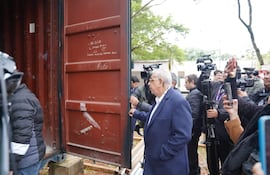 Isaías Fretes, canoso y con abrigo oscuro, revisa un contenedor metálico rojo mientras periodistas observan con atención.
