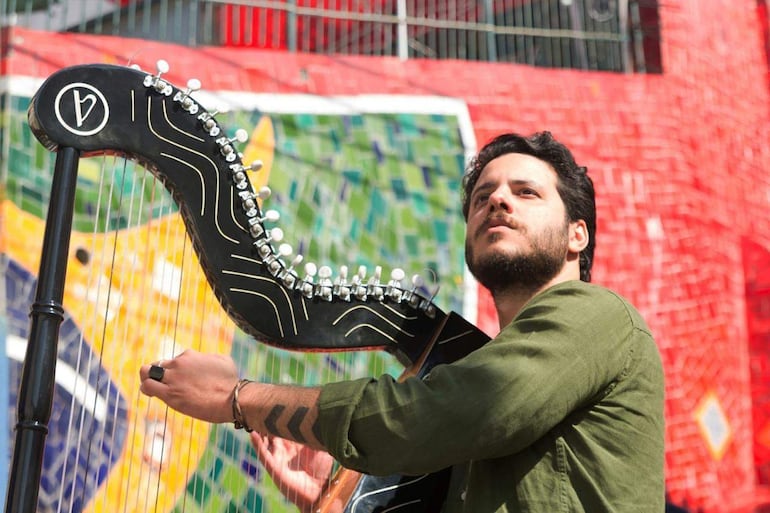 Hombre con cabello oscuro y barba, vestido de verde, tocando un arpa con atención en fondo de mosaicos cálidos.