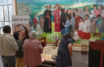 Seis personas, incluidas mujeres y hombres, rezan junto a la tumba iluminada con velas y flores, con mural al fondo.