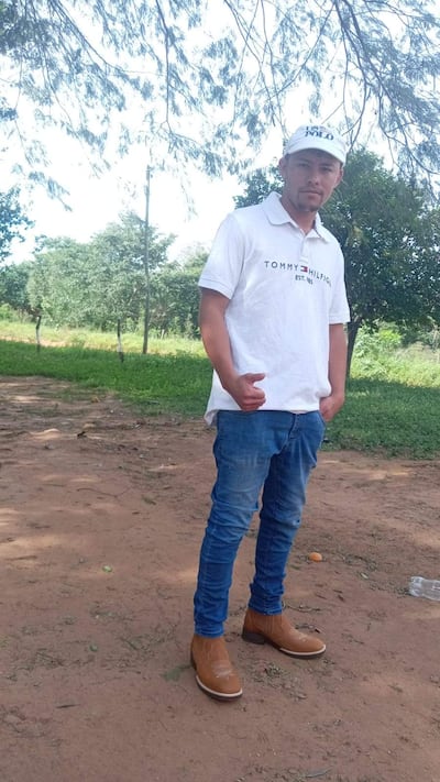 Hombre joven de pie en ambiente rural, lleva camiseta blanca de Tommy Hilfiger y jeans, con expresión tranquila y gesto de pulgar arriba.