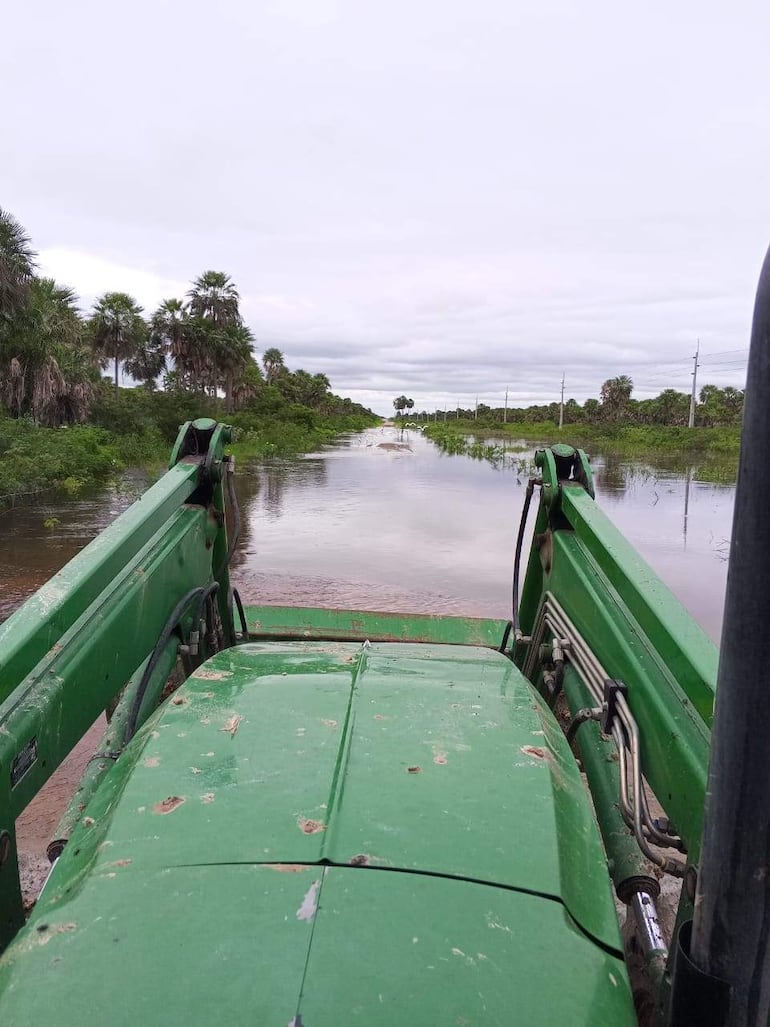 Tractor verde en vía inundada, rodeado de árboles y vegetación, bajo un cielo nublado.