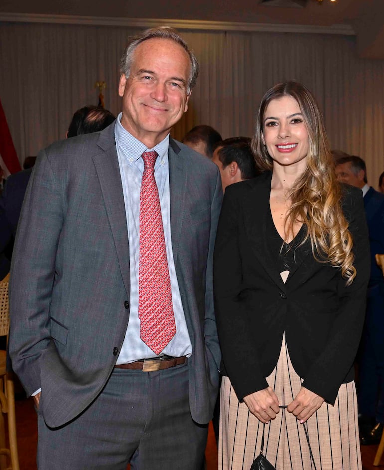 Estefanía Elicetche en blusa negra y falda de rayas, sonríe junto a un hombre en traje gris y corbata roja en un ambiente formal.