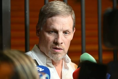 Hombre con cabello corto y barba, vestido con camisa blanca, rodeado de micrófonos y mostrando seriedad.