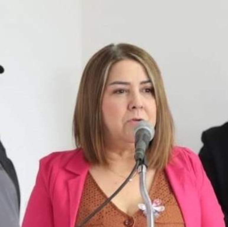 Mujer de cabello castaño y lacio, con blazer rosa, habla ante un micrófono; fondo neutro y figuras indistintas de audiencia.