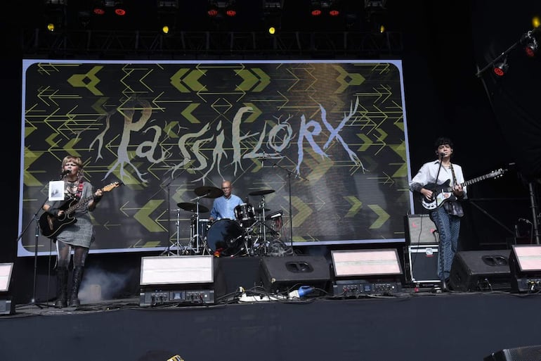 Tres músicos en el escenario, con guitarra y batería, bajo luces brillantes, transmitiendo energía al público.
