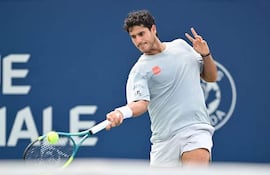 Hombre joven con cabello rizado y camiseta clara golpea una pelota de tenis en cancha con panel azul de fondo.