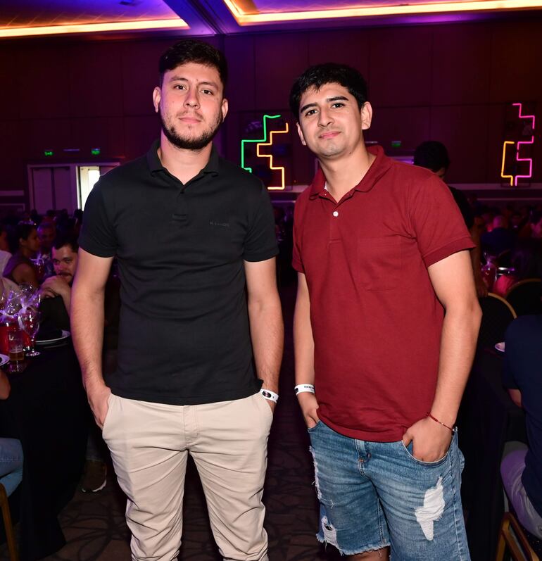 Dos hombres sonríen, uno con camiseta negra y otro con camiseta roja, en un ambiente festivo con luces coloridas.