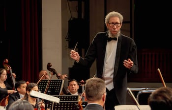 León Burke III, con frac y batuta, dirige a la Orquesta Sinfónica. Músicos en trajes formales, concentrados en el escenario iluminado.
