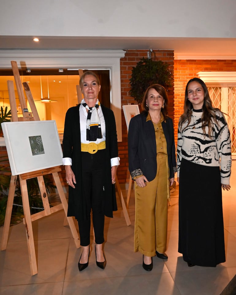 Tres mujeres conversan en un elegante espacio de arte; una viste blusa blanca y abrigo negro, otra traje amarillo y chaqueta oscura, y otra suéter negro.