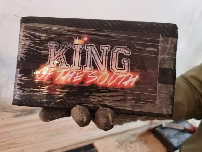 Mano sostiene un paquete envuelto con la leyenda 'KING OF THE SOUTH' y llamas en un fondo neutro.