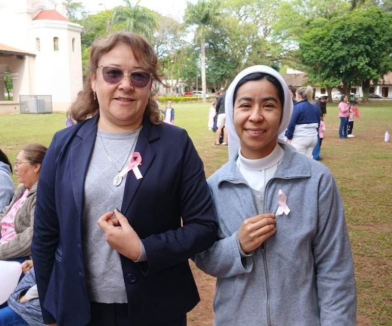 Carolina Flores sonríe con chaqueta azul y blusa gris, junto a la hermana Carmen Alonzo en abrigo gris, sosteniendo cintas rosas en parque.