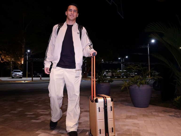 Hombre joven con chaqueta clara y pantalones deportivos empuja maleta dorada, sonriendo en un entorno nocturno iluminado.