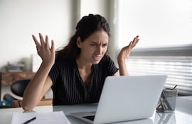 Mujer con cabello largo y oscuro, blusa de rayas, gesticulando frustrada frente a una laptop en una mesa ordenada.