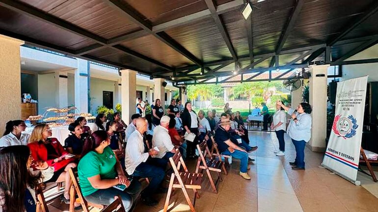Dos mujeres hablando al público, con personas sentadas en sillas de madera y mesas decoradas con plantas y alimentos.