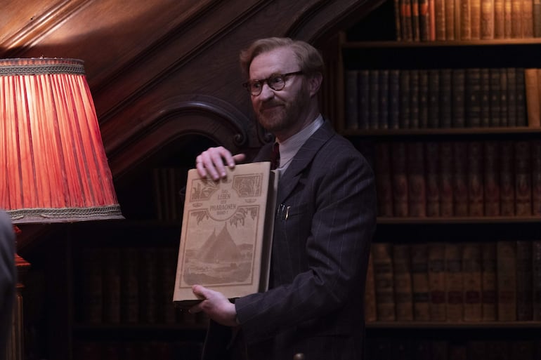 Mark Gatiss de pie con cabello claro y barba, vestido con traje de rayas, presenta un libro sobre pirámides bajo luz cálida.