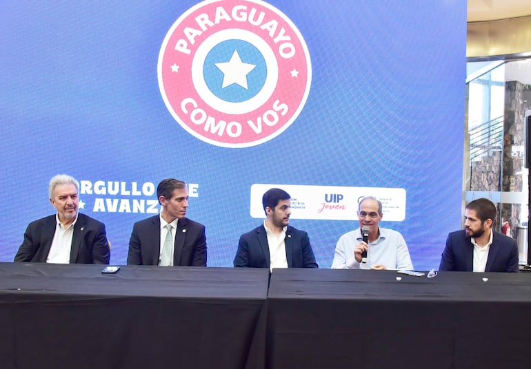Cinco hombres en mesa con mantel negro; uno habla con micrófono, todos visten trajes oscuros y camisas claras. Fondo azul con texto y logotipos.