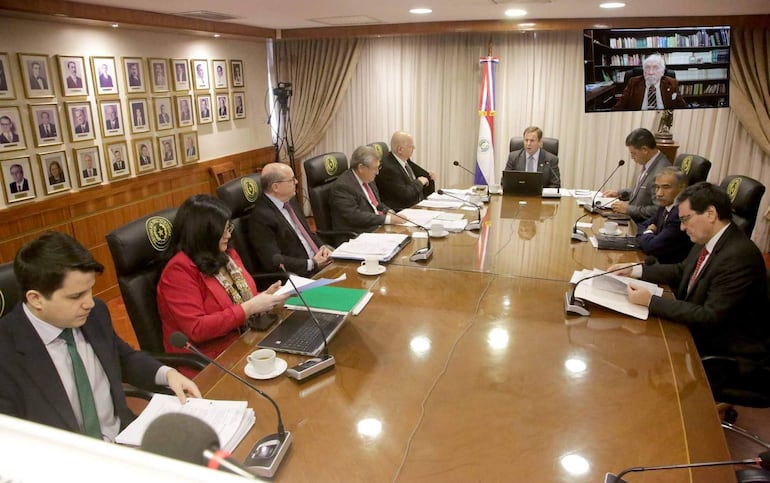 Hombres y mujeres en traje de oficina, reunidos alrededor de una mesa ovalada, con banderas y retratos en un ambiente formal.
