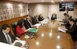 Hombres y mujeres en traje de oficina, reunidos alrededor de una mesa ovalada, con banderas y retratos en un ambiente formal.