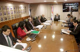 Hombres y mujeres en traje de oficina, reunidos alrededor de una mesa ovalada, con banderas y retratos en un ambiente formal.