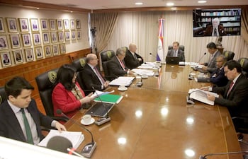 Hombres y mujeres en traje de oficina, reunidos alrededor de una mesa ovalada, con banderas y retratos en un ambiente formal.