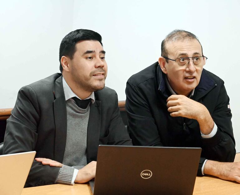 Estigarribia en traje oscuro y Galeano Segovia en chaqueta negra discuten en mesa de madera con laptops abiertas.