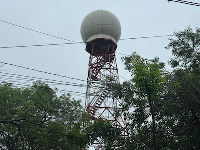 Torre de comunicación alta y metálica, con esfera blanca en su cima, rodeada de árboles y cables eléctricos.