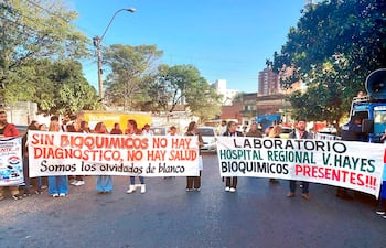 Manifestantes sosteniendo pancartas en demanda de bioquímicos, algunos vistiendo camisetas blancas, en un ambiente urbano.