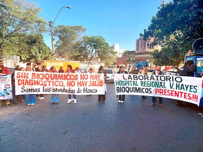 Manifestantes sosteniendo pancartas en demanda de bioquímicos, algunos vistiendo camisetas blancas, en un ambiente urbano.