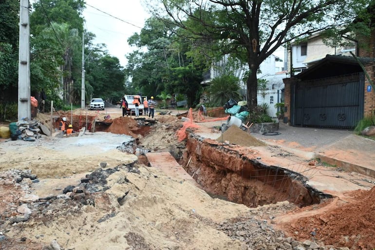 Cinco trabajadores con chalecos reflectantes observan una excavación. Árboles y viviendas se ven al fondo.
