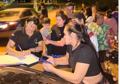 Mujer escribe en el capó de un auto, mientras otra mira su teléfono. Una mujer sostiene a un niño pequeño en un entorno urbano.