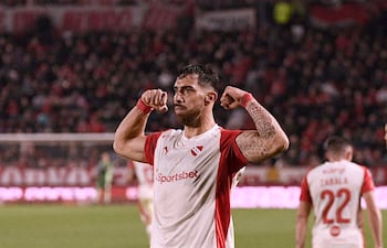 Jugador de Independiente con camiseta blanca y roja en actitud celebratoria; otros jugadores y un público animado en el fondo.