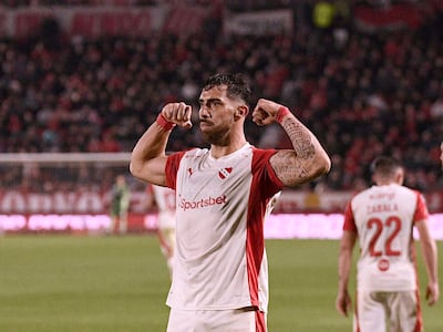 Jugador de Independiente con camiseta blanca y roja en actitud celebratoria; otros jugadores y un público animado en el fondo.