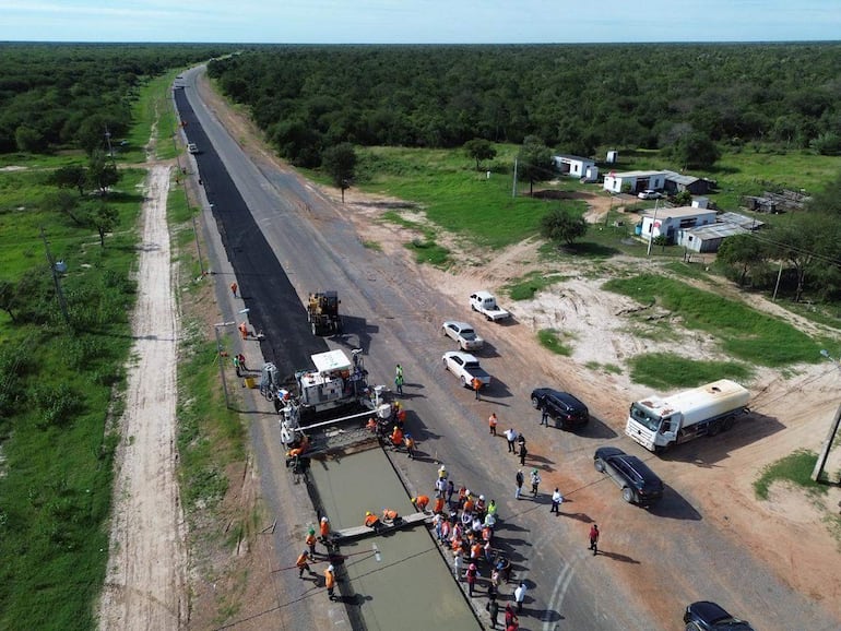 Varios trabajadores con chalecos naranjas operan maquinaria pesada en la construcción de la ruta, rodeados de vegetación y casas modestas.