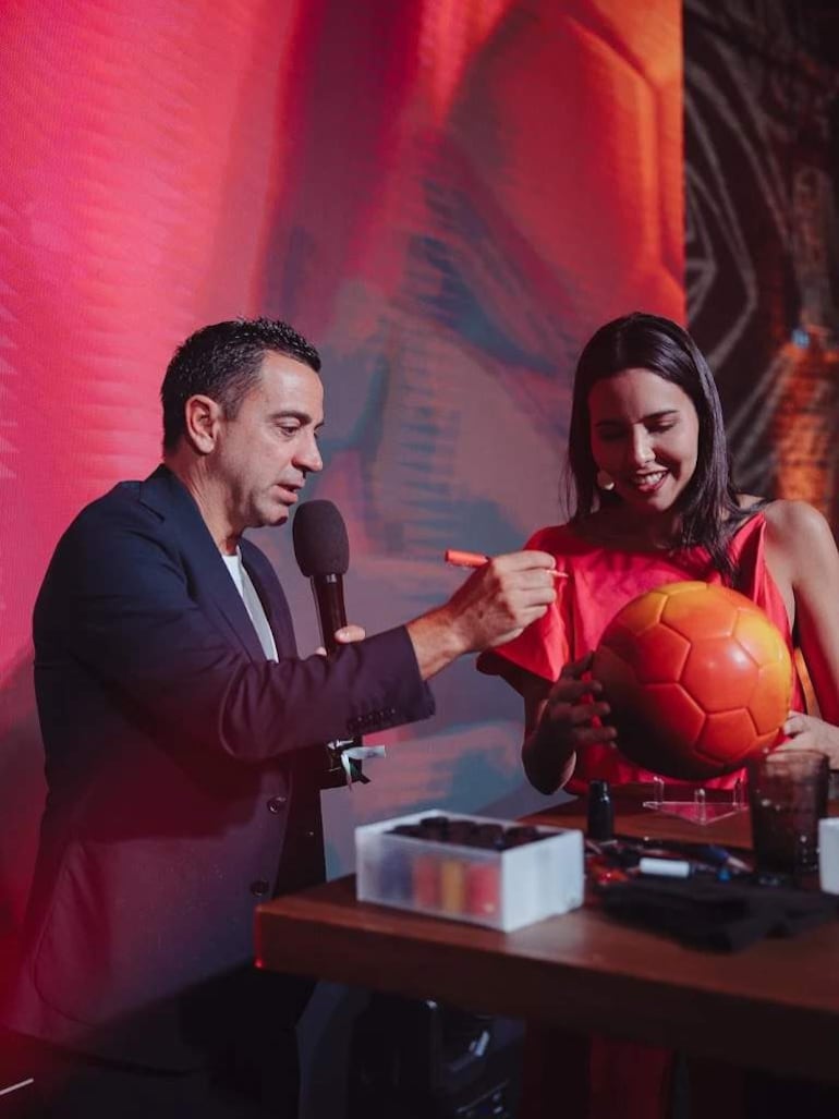 Xavi Hernández habla con micrófono mientras Lili Cantero sostiene un balón de fútbol, ambos en ambiente cálido y festivo.