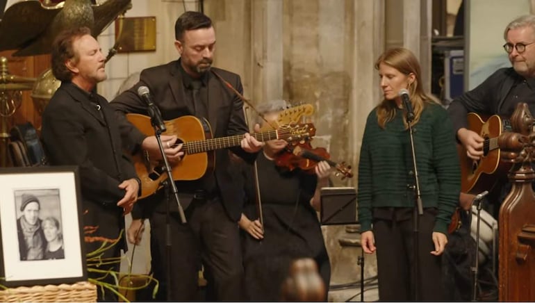 Cuatro personas en escena: hombre con guitarra, mujer con micrófono, homenajeando a Glen Hansard en un ambiente íntimo y formal.
