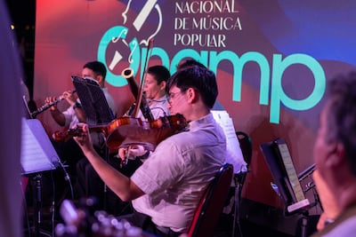 Cuatro músicos de la Orquesta Nacional de Música Popular, todos con camisas blancas, tocan instrumentos en un ambiente iluminado.
