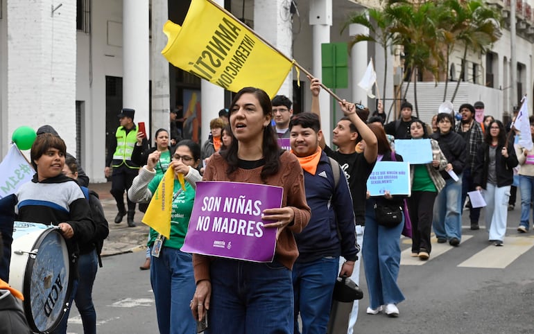 Mujer con cartel 'SON NIÑAS NO MADRES', niño con tambor, otros participantes sosteniendo carteles y banderas en un ambiente urbano.