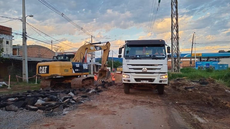 Excavadora amarilla CAT opera en obra de pavimentación, con camión CAT estacionado en el fondo y cielo anaranjado.