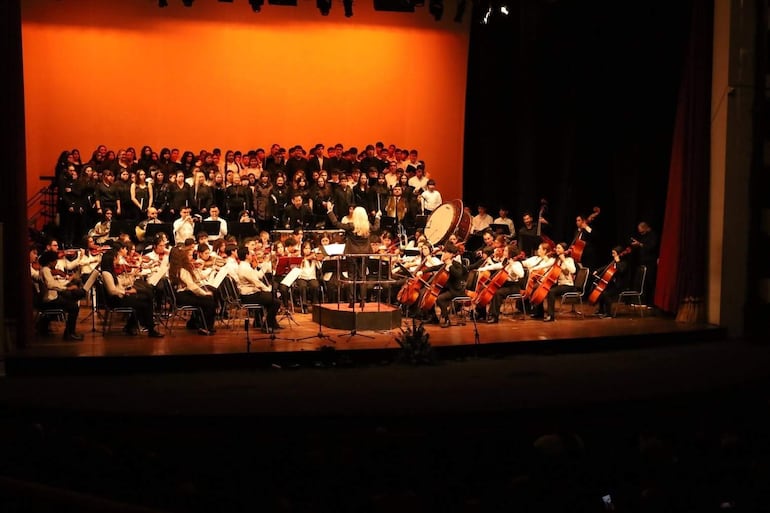 Directora de orquesta rubia y vestida de negro dirigiendo a jóvenes músicos con cuerdas y un coro en un escenario iluminado.