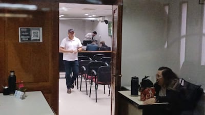 Hombre con camiseta blanca y gorra habla en sala de reuniones, mientras mujer con gafas escucha atentamente, rodeados de más personas.