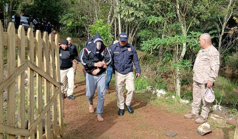 Policías en chalecos oscuros sostienen a Feliciano Martínez Peña, cubierto con bolsa negra, en un entorno boscoso.