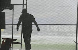 Persona con ropa oscura en movimiento bajo intensa lluvia, con valla metálica en el fondo y entorno deportivo difuso.