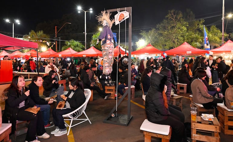 Multitud de personas sentadas en bancos de madera en un evento al aire libre, con carpas rojas y luces decorativas detrás.