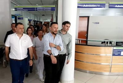 Richard Ramírez en camisa clara habla con autoridad en hospital, mientras Derlis León observa atento, entorno profesional con personal médico.
