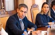 Marco Riquelme, en traje oscuro, y Mónica Recalde, en abrigo oscuro, conversan en mesa de madera durante reunión formal.