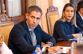 Marco Riquelme, en traje oscuro, y Mónica Recalde, en abrigo oscuro, conversan en mesa de madera durante reunión formal.