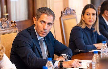 Marco Riquelme, en traje oscuro, y Mónica Recalde, en abrigo oscuro, conversan en mesa de madera durante reunión formal.