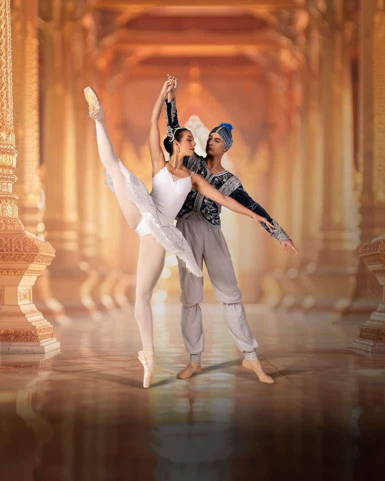 Bailarines en escena: mujer en tutu blanco y hombre en atuendo azul y gris, sosteniéndose de las manos en un entorno palaciego.